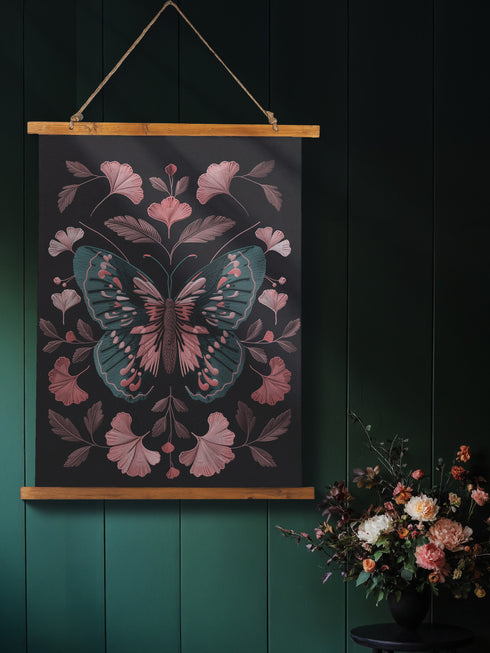 Hanging tapestry featuring a teal and pink butterfly surrounded by ginkgo-like leaves on a black background. The textile is suspended from wooden dowels with twine, creating detailed 3D embroidery-like texture.